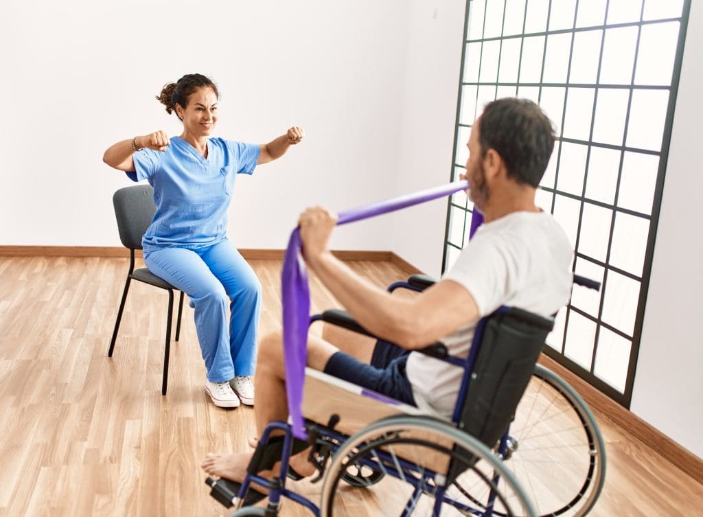 hombre y mujer de mediana edad en sesion de rehabilitacion usando banda elastica sentado en silla de ruedas en la clinica de fisioterapia scaled